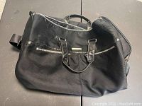 Black Versace Parfums travel bag with leather trim, two top handles, zipper closure, and detachable shoulder strap sitting on a flat surface.