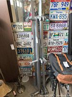 Adjustable metal bed frame leaning against a wall with vintage license plates behind it, showing the lower section including casters and the adjustable side rails.