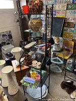 Front and side view of metal cabinet with glass shelves filled with various household items including ceramics, glassware, and decor items.