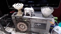 Photo showing a metal decorative clock, elephant figurine, punch bowl with cups, and compote on a black surface