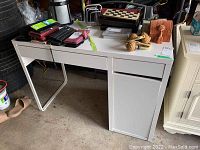 Front view of white desk with two drawers and a metal support leg beneath, surface has some marks and miscellaneous items on top.