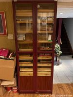 Full view of tall wooden humidor cabinet with two glass doors and wooden shelves inside filled with various cigar boxes and tubes.