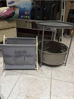 Image showing a round grey metal table with tripod legs and fabric basket underneath next to a fabric magazine rack with wooden frame and three front pockets on tiled floor.