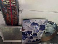 Lot showing framed canvas print with blue and white floral design, square mirror with white frame and round mirror with wooden frame stacked against a wall.