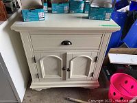 Front view of off-white wooden nightstand with drawer and lower cabinet showing metal hardware and scratches.