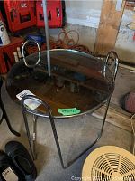 Metal side table with round mirrored top, some marks visible on the mirrored surface. Table has three metal legs with curved handles on opposite sides.