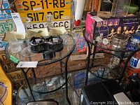 Two metal side tables with glass tops and decorative black metal floral designs on frames in a garage setting with various items around