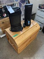 Wooden toy trunk with two black Honeywell fans placed on top. The trunk is rectangular with rope handles and a natural wood finish.