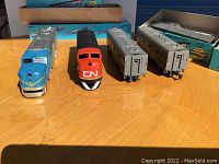 Five Athearn model train pieces including locomotives in various railroad liveries and two silver train cars, displayed outside on wood tabletop with blue storage doors in background.