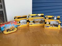 Five boxed CAT branded die-cast model construction vehicles displayed on a table, showing packaging and visible models.