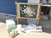 Overview of entire lot showing floral fabric in wooden frame, small storage drawers, basket with crafting tools, multiple embroidery floss skeins, and various kits and fabrics laid out on ground.