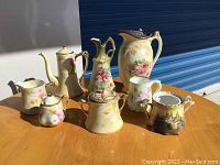 Full set of vintage porcelain china items on wooden table under sunlight, showing various pitchers, creamers, sugar bowls, and decorative vases with floral and landscape patterns.