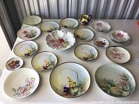 Multiple porcelain plates and a serving bowl arranged on a table showing various fruit and flower hand-painted designs. Includes different sizes and styles. Condition shows usage wear.