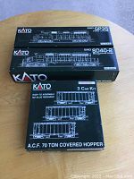 Three Kato Precision Railroad Models boxes stacked on table: two locomotive model boxes (EMD GP35 and EMD SD40-2) and one 3-car kit box of A.C.F. 70 Ton Covered Hopper cars.
