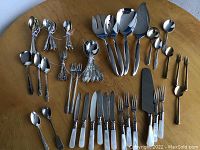 Mixed flatware including multiple sets of spoons, forks, knives, and serving utensils laid out on a wooden table for condition and quantity assessment.