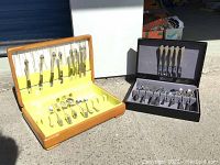 Two flatware sets shown in their display cases on pavement under sunlight, one wooden case with yellow lining and the other black case with gray lining