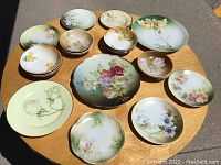 Array of 15 floral china plates and bowls arranged on a wooden table, displaying variety of floral designs and color schemes in pastel shades.