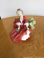 Front view of Royal Doulton Lydia figurine showing lady in red dress and hat sitting among green foliage on wooden surface.