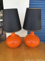 Pair of retro orange table lamps with rounded bases and black lampshades on a wooden surface, showing overall condition and size.