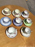 Photo showing 8 sets of assorted floral and colored tea cups and saucers on a wooden table.