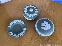 Three green Jasperware pieces on wood table: a plate, a small lidded box with vine relief, and a larger lidded box with chariot scene.