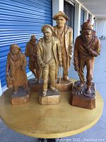 Group of six detailed wood carvings displayed on a round wooden table, showing full figures of men in various clothing and accessories.