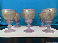 Photo showing 6 purple and beige handpainted glass goblets with drip and textured patterns, each with a gold sticker, arranged on a white base with a blue background.