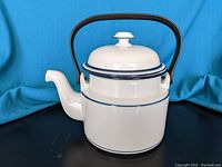 White Dansk Bistro tea pot with blue bands, black handle, on black surface with blue backdrop