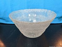 Clear textured glass bowl with flared top shown from angle, against blue fabric back and black surface.