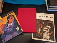 Three art books side by side on a black surface. Left book is Gauguin with illustrated cover featuring a woman holding a tambourine, middle book is a red cloth hardcover with no visible text, right book is André Derain with still life painting cover.