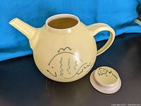 Large round ceramic teapot in a creamy yellow color with a simple fish motif, viewed from the side showing the spout and handle with lid off.