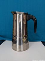 Front view of stainless steel mocha espresso maker with black handle against blue background, showing entire coffee maker including spout.