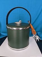 Front view of green metal ice bucket with silver lid topped with duck figure and attached ice tongs