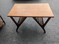 Front angled view of vintage mid century coffee table showing rectangular wooden top with some wear and unique angled wooden legs with built-in slatted shelf below.