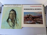Two art books laying flat; one titled 'History In Their Blood' with a painted Native American portrait on cover, and the other 'Remington & Russell' from the Sid Richardson Collection.