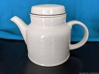 Side view of white ceramic teapot with lid on, showing embossed floral pattern and handle shape.