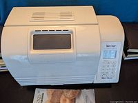 White Westbend bread maker machine with control panel and window on lid, placed on surface with booklet underneath.