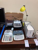Overview of lot including two adding machines, one label maker, APC Back-UPS battery backup, and desk lamp on wooden desk.