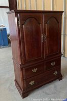Front view of wooden 4-drawer armoire showing closed upper cabinet doors with arched panels, decorative trim on top and base, and metal handles and pulls, with scratches.