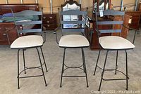Front view of three black metal swivel bar stools with light upholstered seats, two without armrests and one with armrests in the center, showing wear and staining on seats.