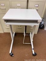 Front view of a white metal and laminate rolling cart on four casters with a lower shelf and a sticker residue on top.