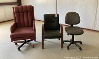 Photo of three chairs including a burgundy upholstered rolling high-back chair, a dark wood framed upholstered chair with fabric upholstery, and a gray fabric rolling desk chair on casters.