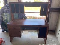 Front view of brown laminate Sauder desk with hutch, drawers on left, open space on right, visible wear on desktop.