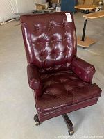 Full view of the burgundy upholstered desk chair with button tufting, wooden wheels, and armrests in a warehouse setting.