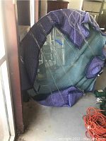 Round glass table top covered partially with blue moving blankets leaning against a wall in a storage area. Shows clear beveled edges and a smooth surface.