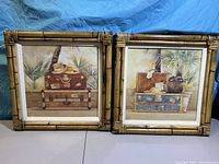 Two bamboo-framed art prints showing artwork of vintage suitcases topped with hats and tropical plants in background.