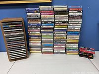 Photo showing stacks of cassette tapes arranged vertically with a wooden storage case holding several tapes on left side, three small black and red boxes on right side.
