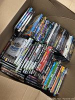 Top view of a large cardboard box filled with DVDs and Xbox 360 games, showing titles and cases stacked closely together.