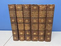 Front view of six hardcover books showing spines with gold embossed titles and volume information. Volumes 1-5 and 7 present.