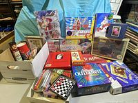 Image showing large assortment of boxed board games, puzzles, and card games on table surface including Scrabble, Scattergories, Monopoly Millennials, Santa puzzle, and Springbok Kids puzzle.
